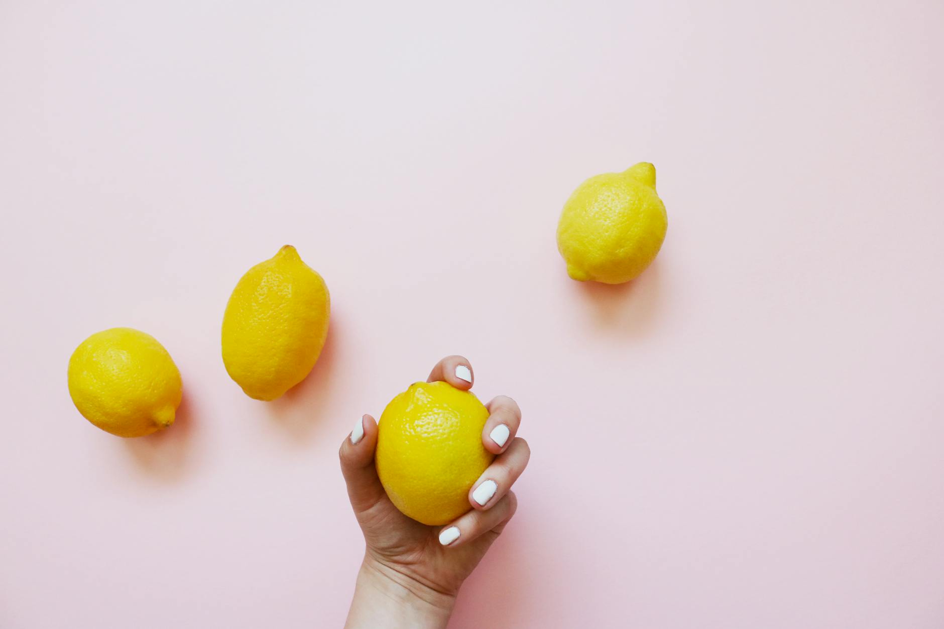 A hand with white nails holding a fresh lemon on a soft pink background