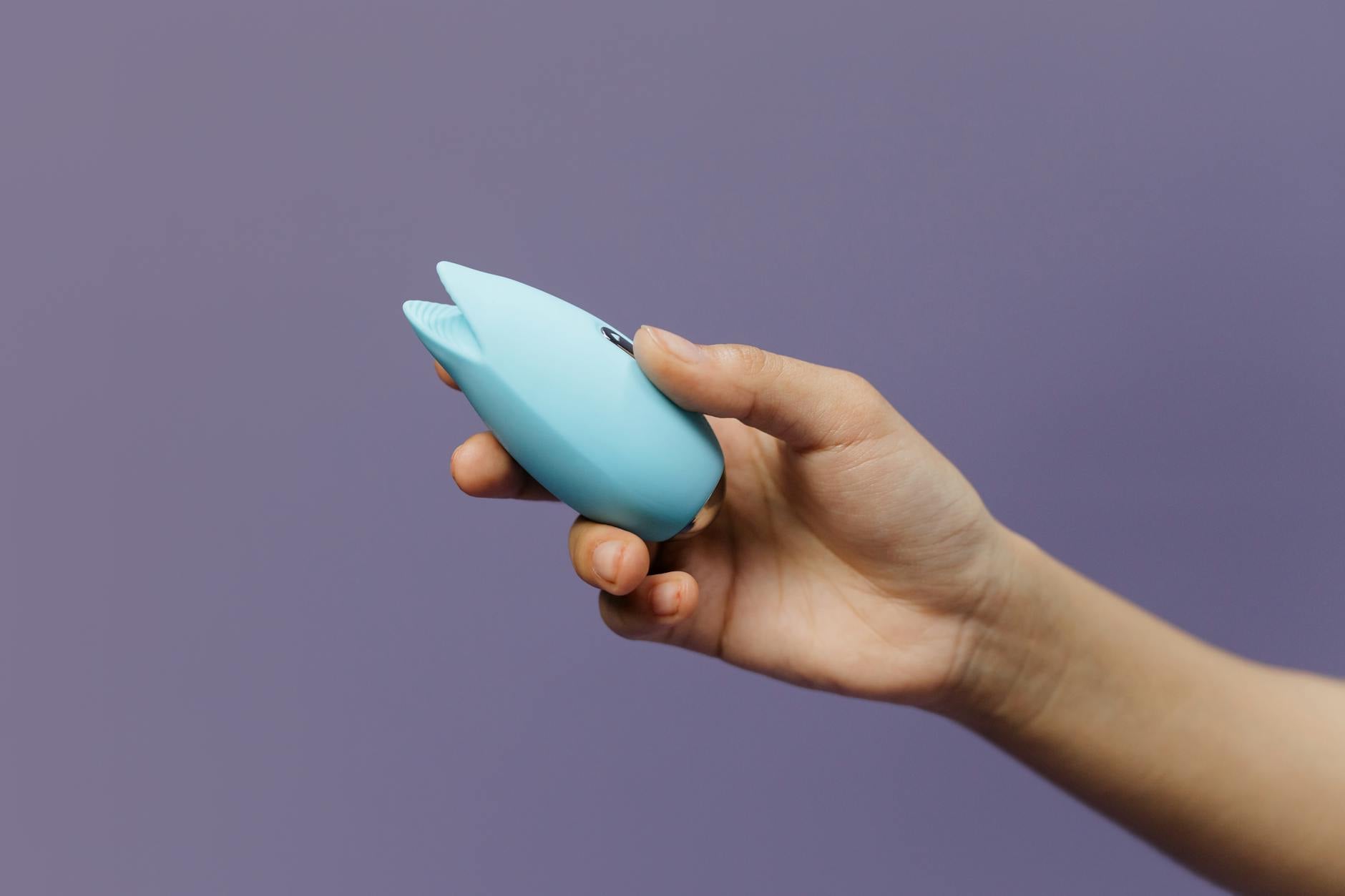 A blue silicone clitoral vibrator held in hand against a purple background, representing tactile pleasure recovery
